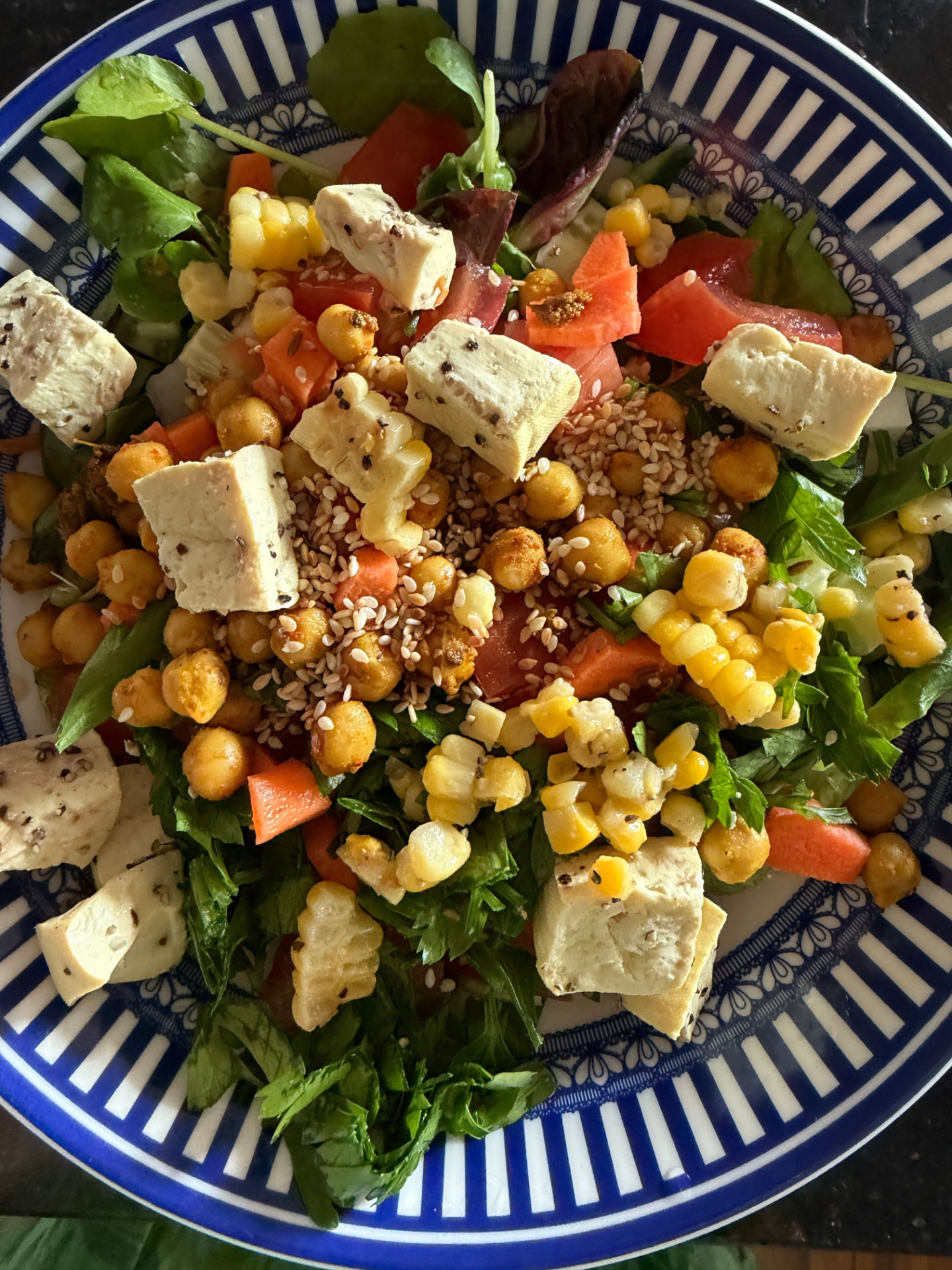 A colorful plant-forward salad with greens, corn, tomatoes, tofu, and chickpeas in a blue-rimmed bowl