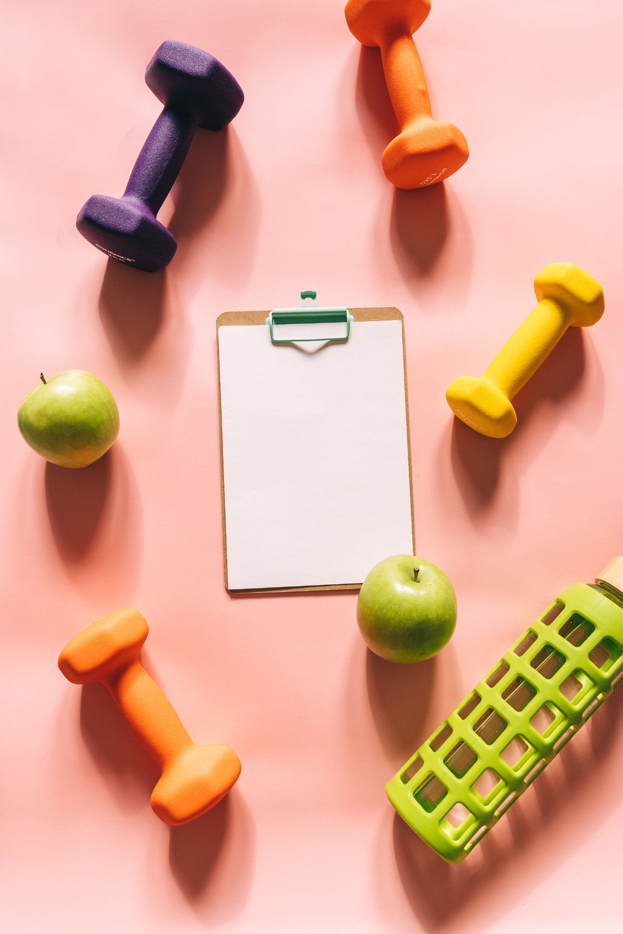 Dumbbells, fresh apples, a water bottle, and a wellness planning clipboard arranged on a soft surface